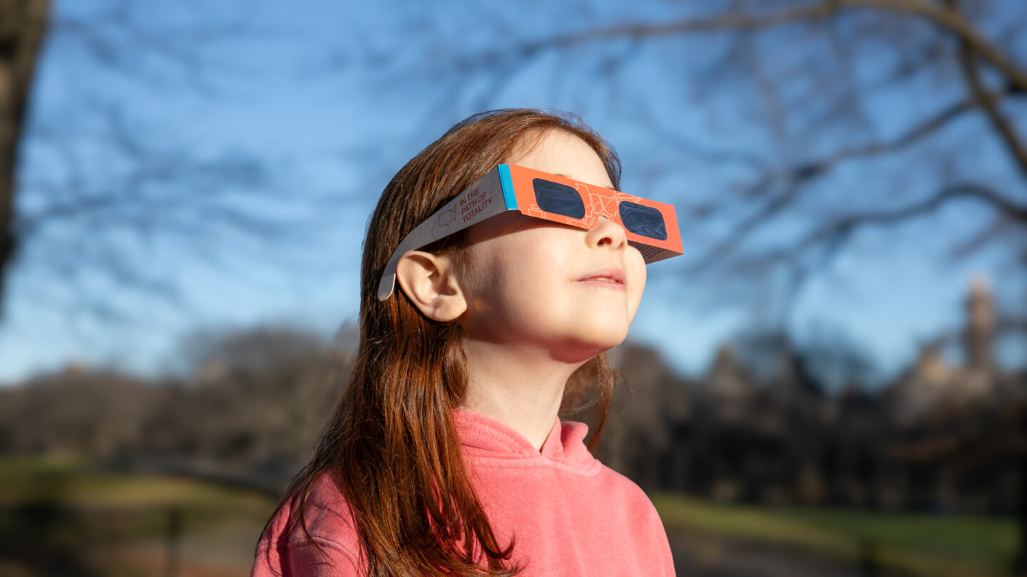 A young girl in a pink hoodie wears solar eclipse glasses and looks up at the sky in a sunlit park with bare trees and blurred buildings in the background.