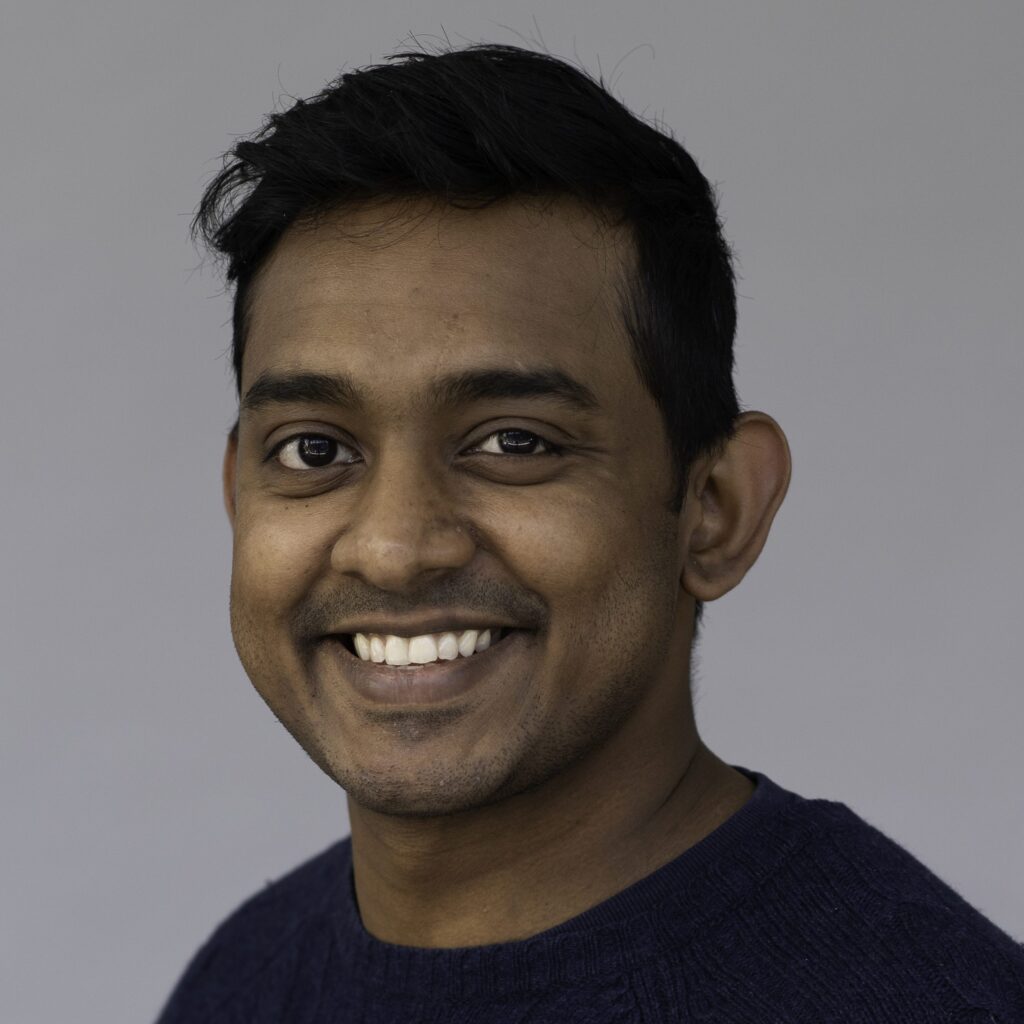 A man with short black hair smiles at the camera. He is wearing a dark blue sweater and standing in front of a plain, light grey background.