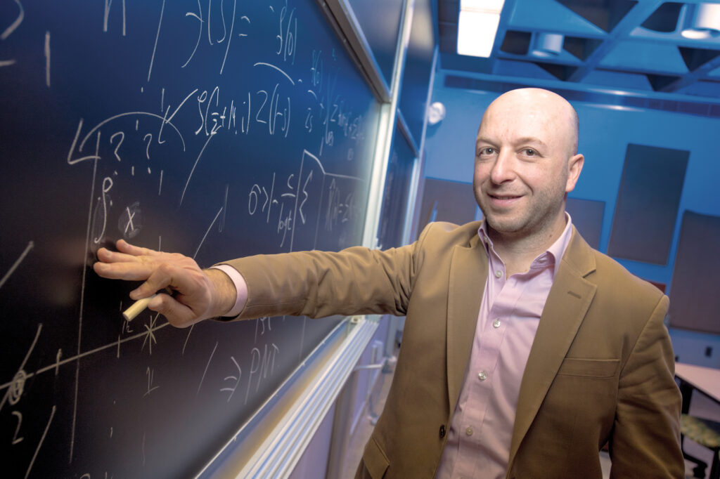 Portrait of Alex Kontorovich writing on a blackboard.