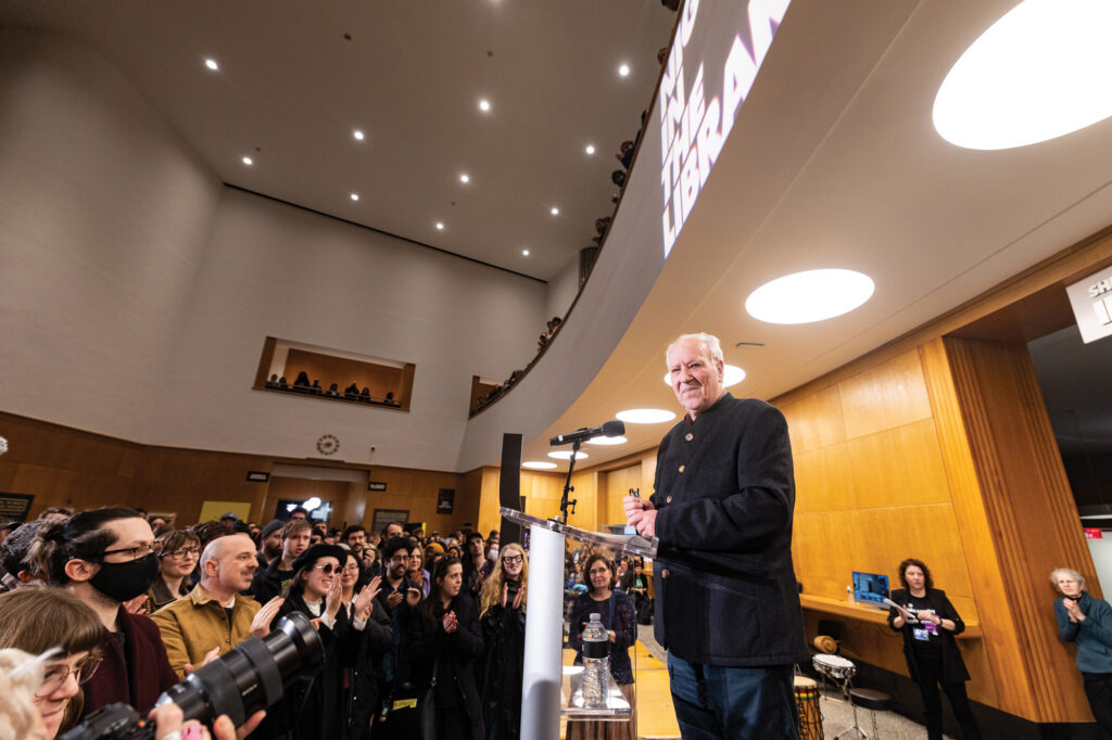 hoto of filmmaker Werner Herzog at a podium speaking to a crowd inside the Brooklyn Library.