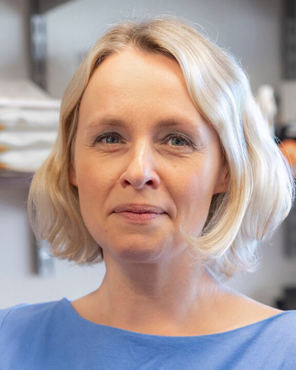 A woman with short blonde hair and a blue top is standing indoors in front of shelves with papers and office supplies, looking slightly toward the camera with a neutral expression.
