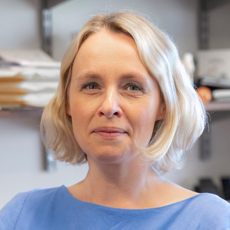 A woman with short blonde hair and a blue top is standing indoors in front of shelves with papers and office supplies, looking slightly toward the camera with a neutral expression.