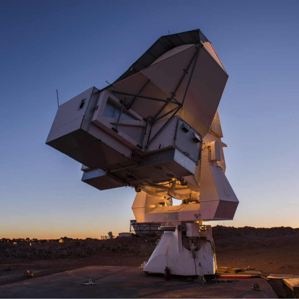 A large radio telescope dish aims toward the sky at sunset, set on a barren, rocky landscape. The sky is clear with a gradient from deep blue to orange near the horizon.