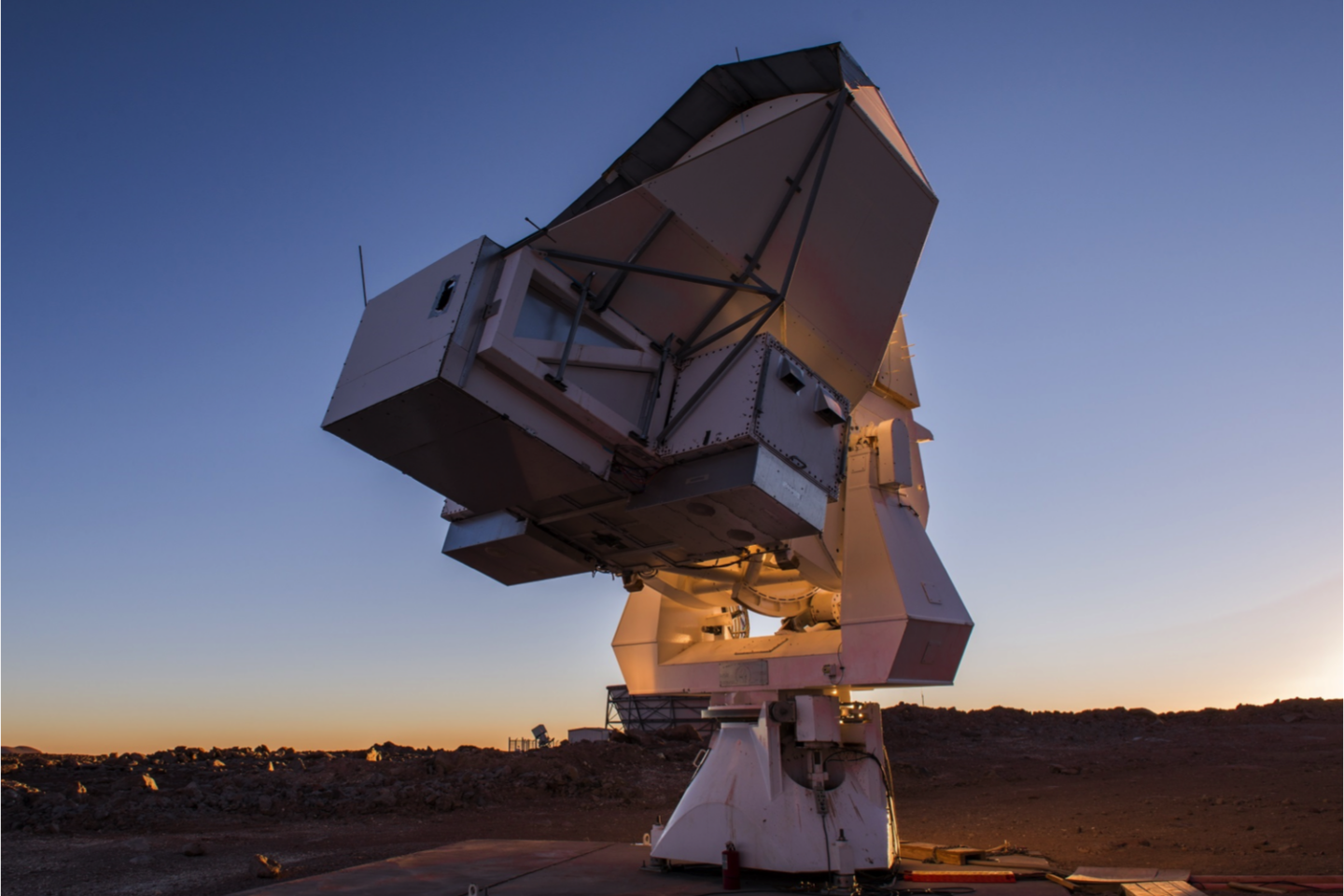 A large radio telescope dish aims toward the sky at sunset, set on a barren, rocky landscape. The sky is clear with a gradient from deep blue to orange near the horizon.