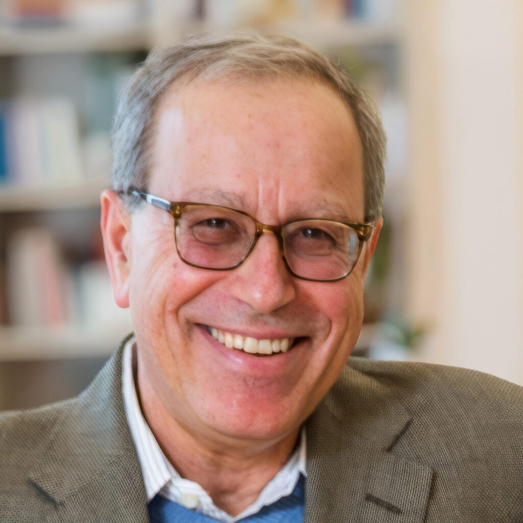 Smiling older man with short gray hair, glasses, and a brown blazer, standing indoors with blurred bookshelves in the background.