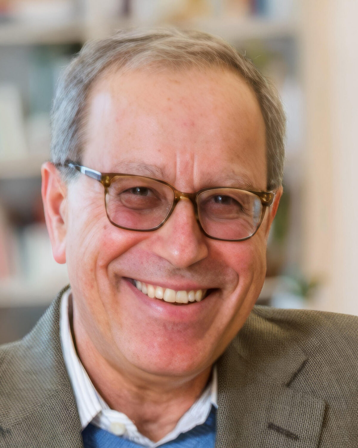 Smiling older man with short gray hair, glasses, and a brown blazer, standing indoors with blurred bookshelves in the background.