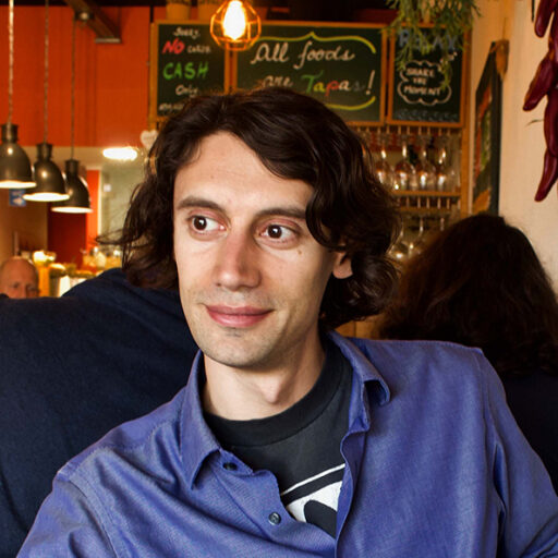 A man with wavy brown hair and a blue shirt sits in a cozy restaurant with chalkboard menus and warm lighting in the background.