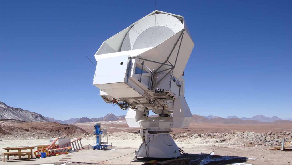 A large white radio telescope with a bowl-shaped dish sits on a concrete platform in a desert, surrounded by mountains under a clear blue sky.