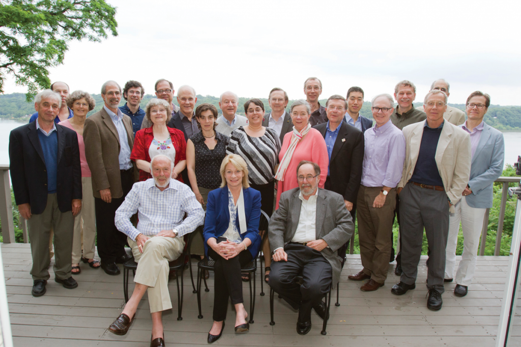 A group of twenty-five adults poses for a photo on a wooden deck with trees and water in the background; three are seated in front while the others stand behind them, all dressed in business or casual attire.
