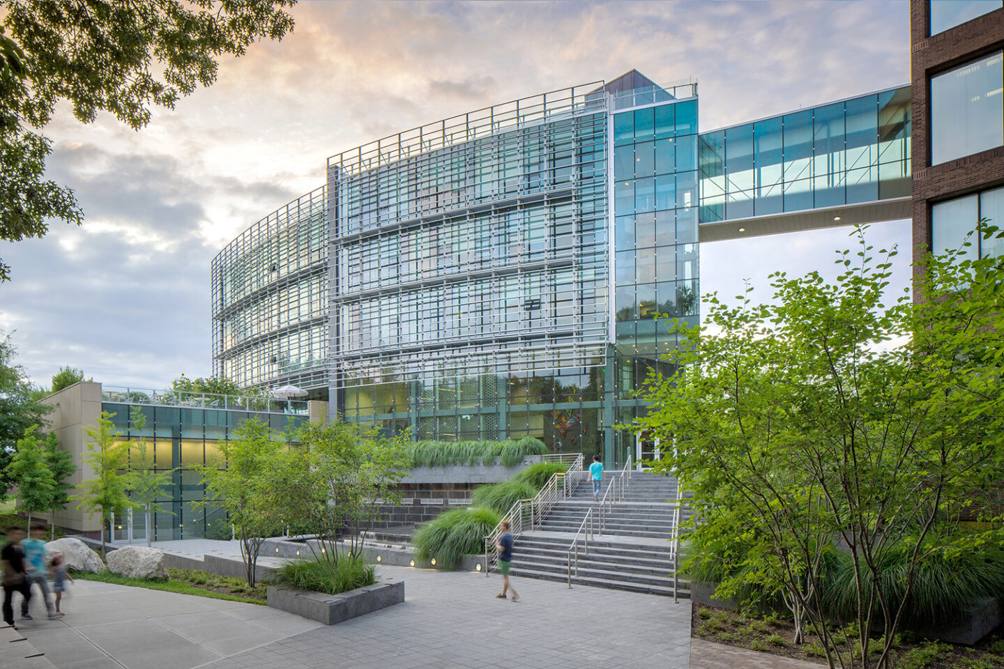 Modern glass-fronted building with multiple stories, a sky bridge, and wide steps leading to the entrance. People walk nearby, and green landscaping surrounds the scene under a partly cloudy sky.