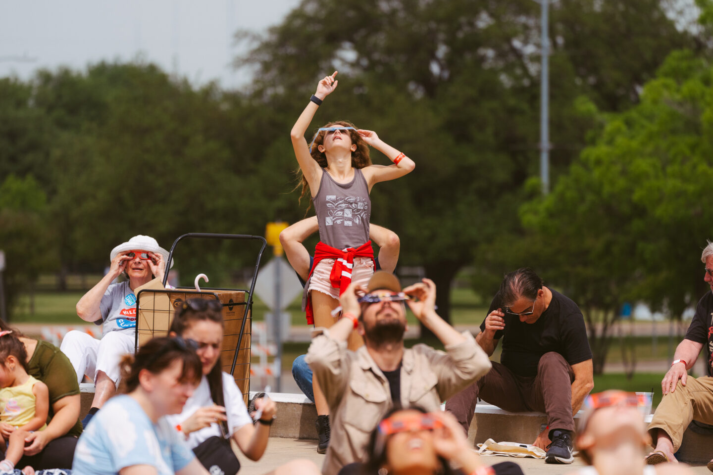 A group of people outdoors watch the sky using solar eclipse glasses. A girl on someones shoulders points upward, while others sit or stand, all focused on the sky. Trees and a park-like setting are visible in the background.