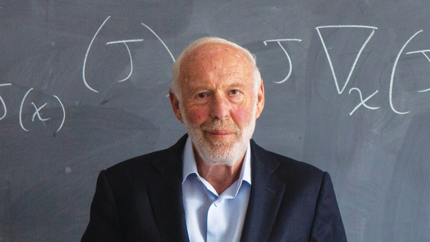Photo of Jim Simons in front of a chalk board that shows an equation.