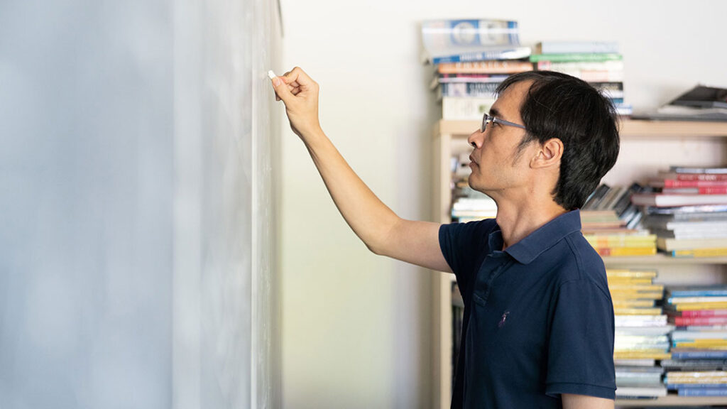 A man wearing glasses writes on a white board next to a bookshelf filled with books.