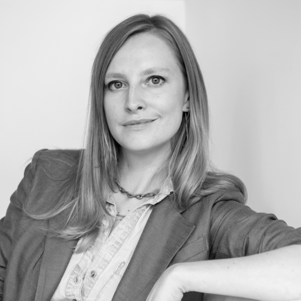 Black and white portrait of a woman with long hair, wearing a blazer and necklace, sitting and looking confidently at the camera with a slight smile.