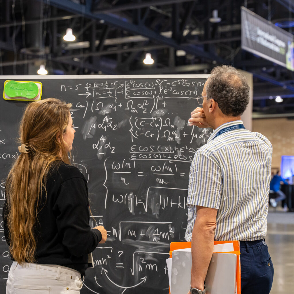 Two people stand in front of a large chalkboard covered in complex math equations at what appears to be a conference or science event in a spacious, modern indoor setting.