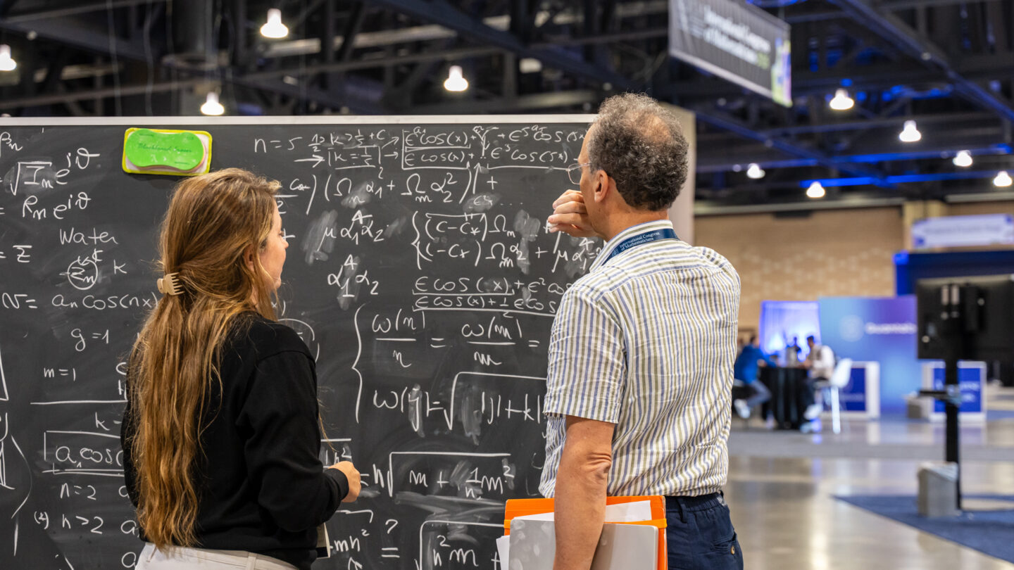 Two people stand in front of a large chalkboard covered in complex math equations at what appears to be a conference or science event in a spacious, modern indoor setting.