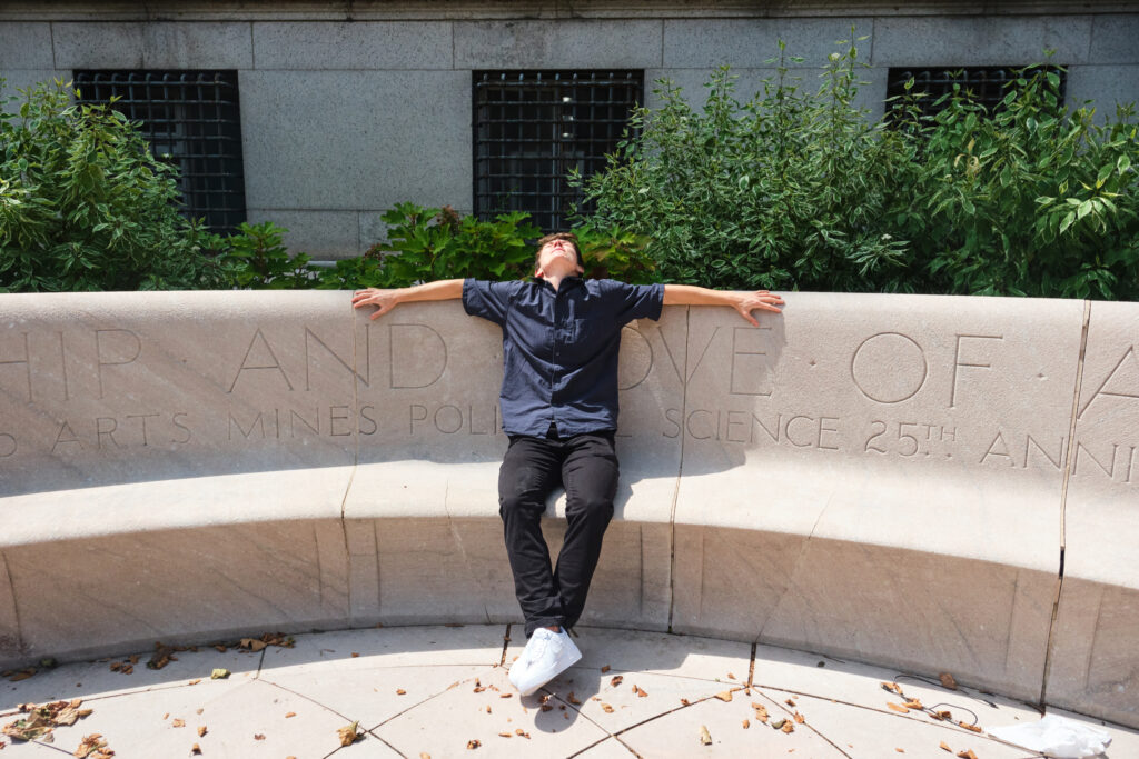 A person in a dark shirt and pants sits on a curved stone bench with arms outstretched, head tilted back, and eyes closed. Green plants and a building with barred windows are in the background.