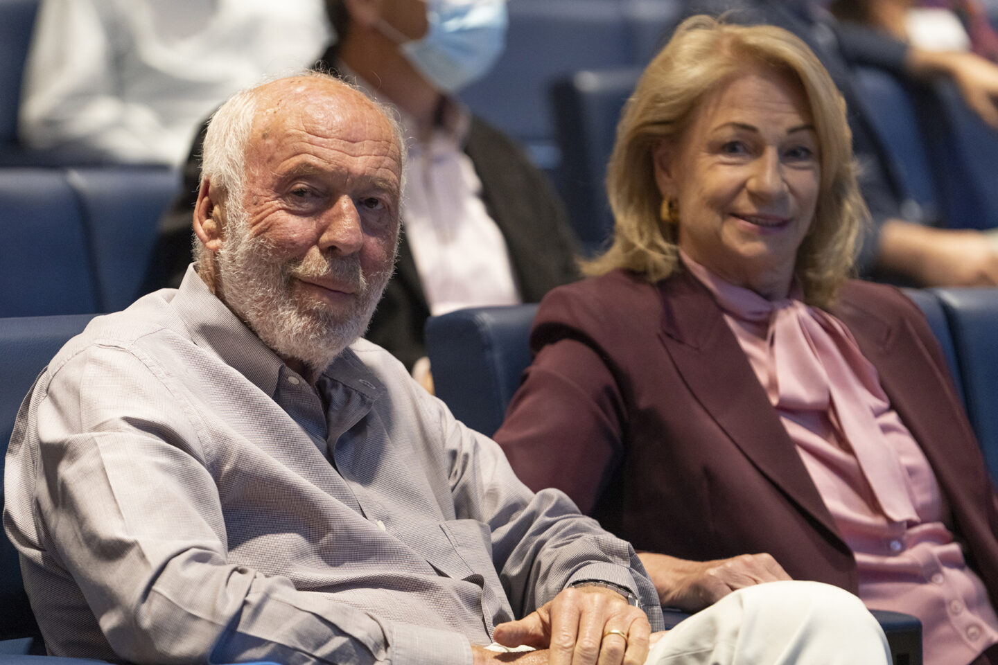 An older man and woman sit next to each other in blue auditorium seats, smiling toward the camera. The man wears a light shirt, and the woman wears a maroon blazer with a pink blouse. Other people are seated behind them.