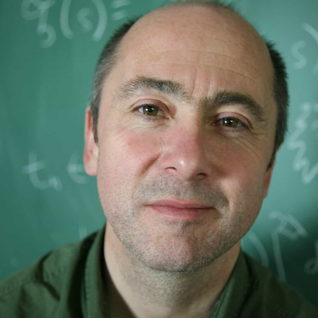 A middle-aged man with a receding hairline stands in front of a green chalkboard covered with mathematical equations, looking slightly up at the camera and smiling gently.