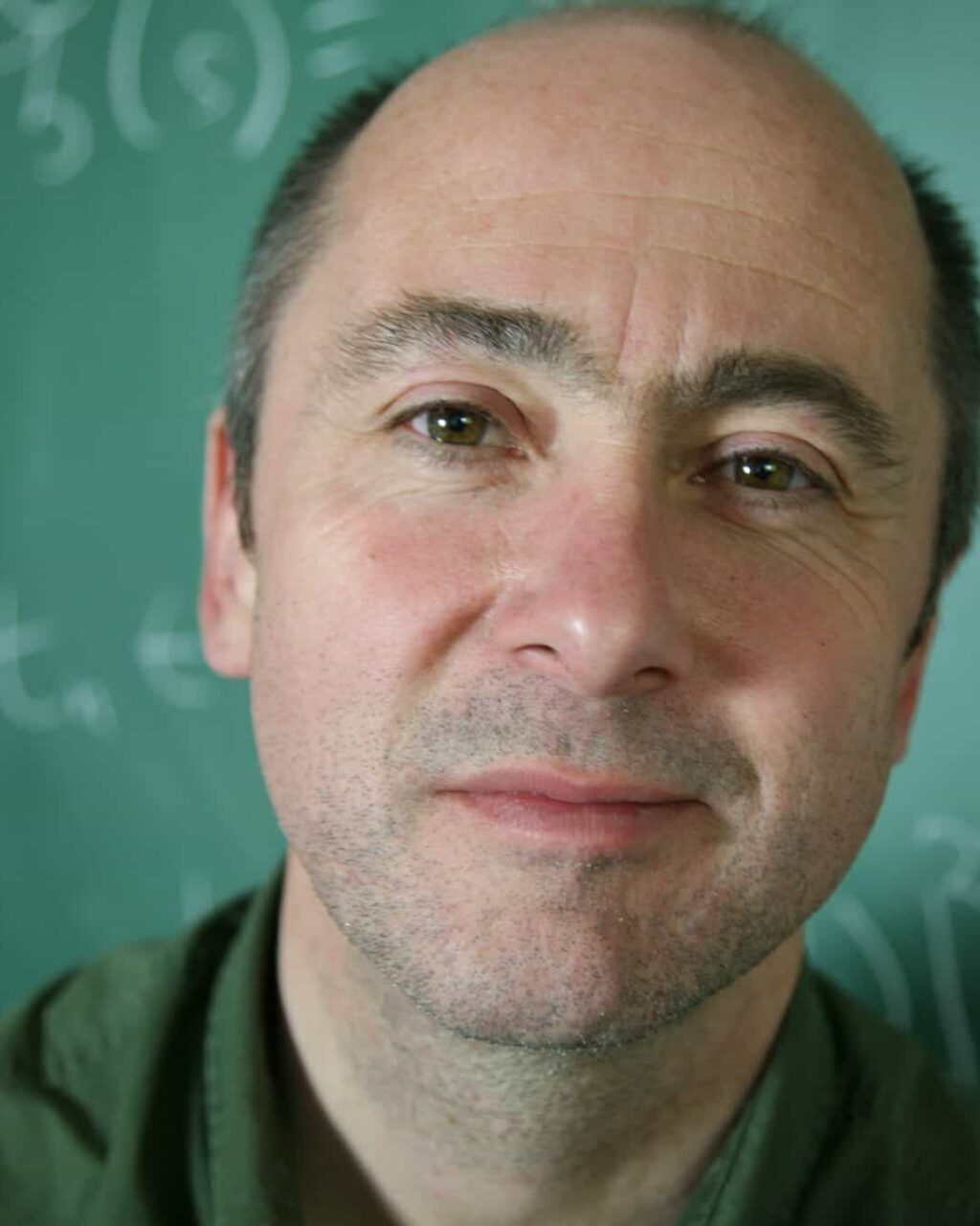 A middle-aged man with a receding hairline stands in front of a green chalkboard covered with mathematical equations, looking slightly up at the camera and smiling gently.