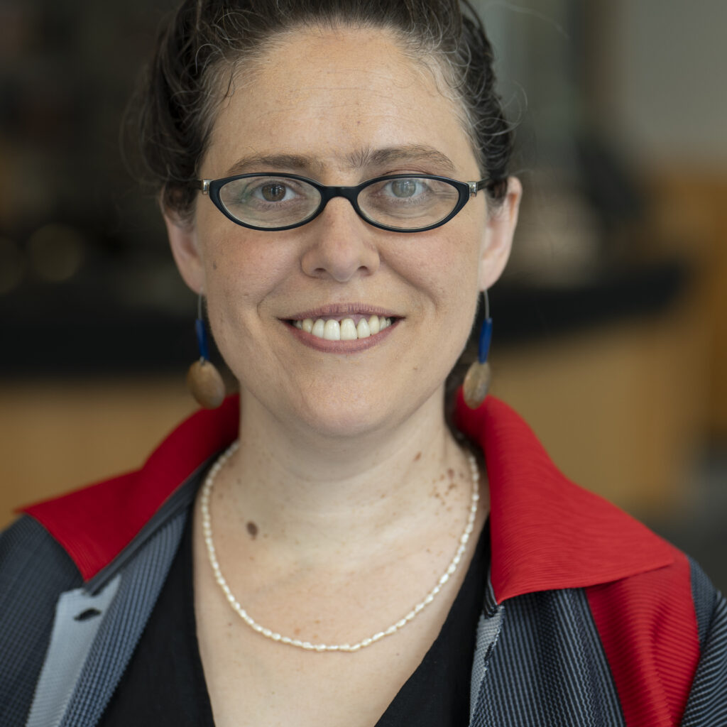 A woman with glasses and curly hair in a bun smiles at the camera. She wears a pearl necklace, blue and red jacket, and dangly earrings. The background is blurred.