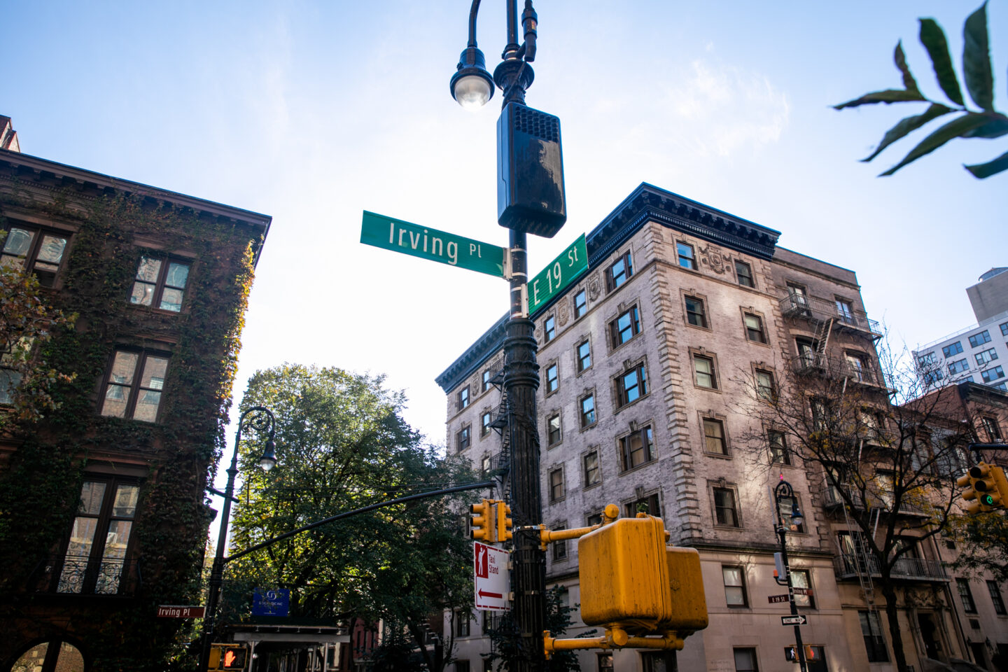 Street signs for Irving Place and E 19th St on a pole with a traffic light, surrounded by old brick buildings, trees, and a clear blue sky.