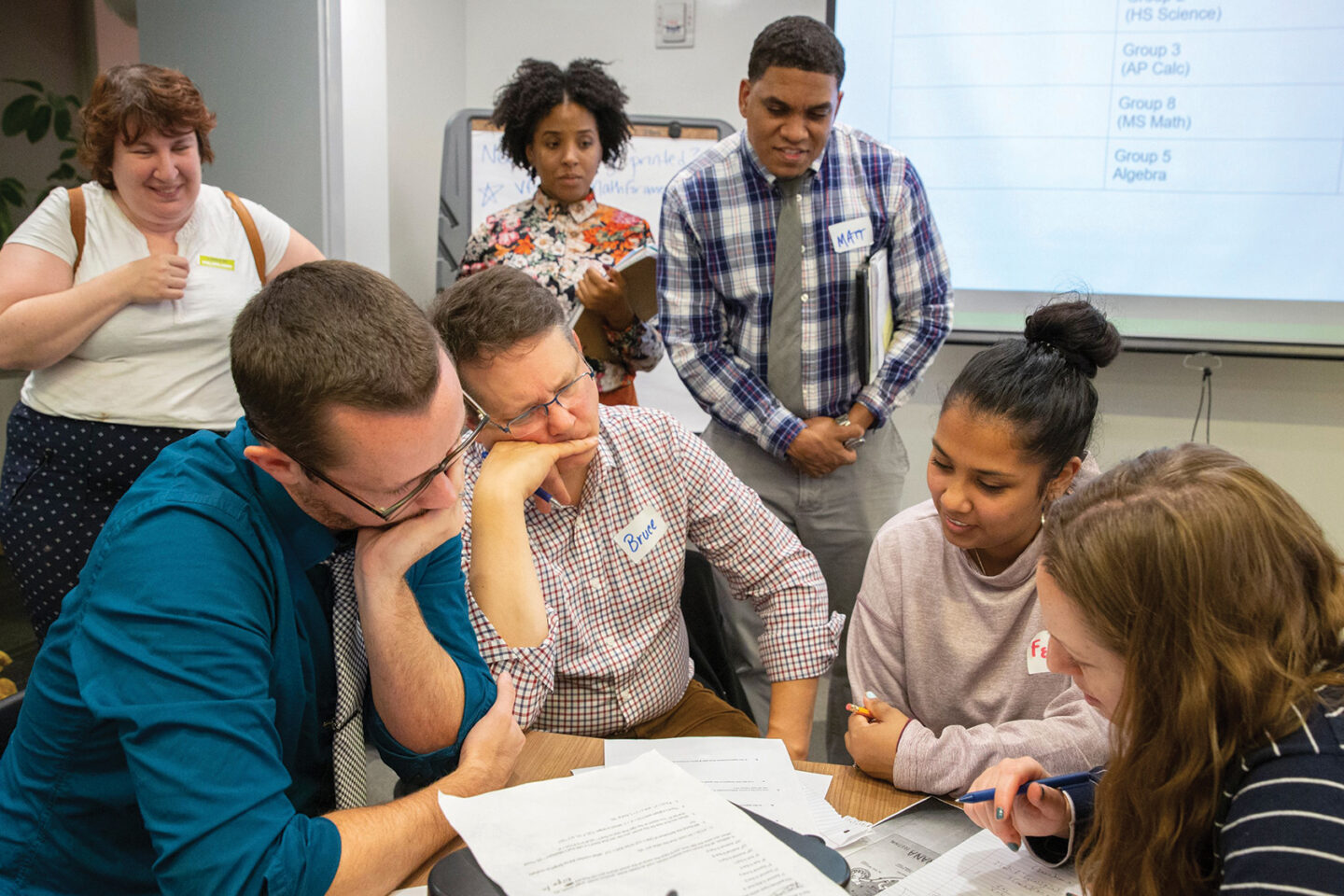 A group of people sits at a table collaborating on paperwork, while others stand behind them observing and smiling. A whiteboard with charts is visible in the background.