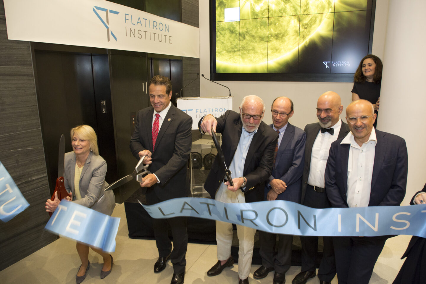 Seven people, dressed in business attire, smile and cut a ceremonial ribbon at an indoor event. A Flatiron Institute sign and a large sun image are visible in the background. One woman and six men participate in the ceremony.