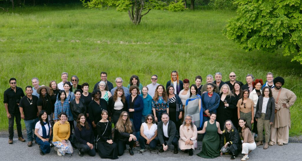 A large group of people pose together outdoors on a paved area in front of green grass and trees, smiling at the camera.