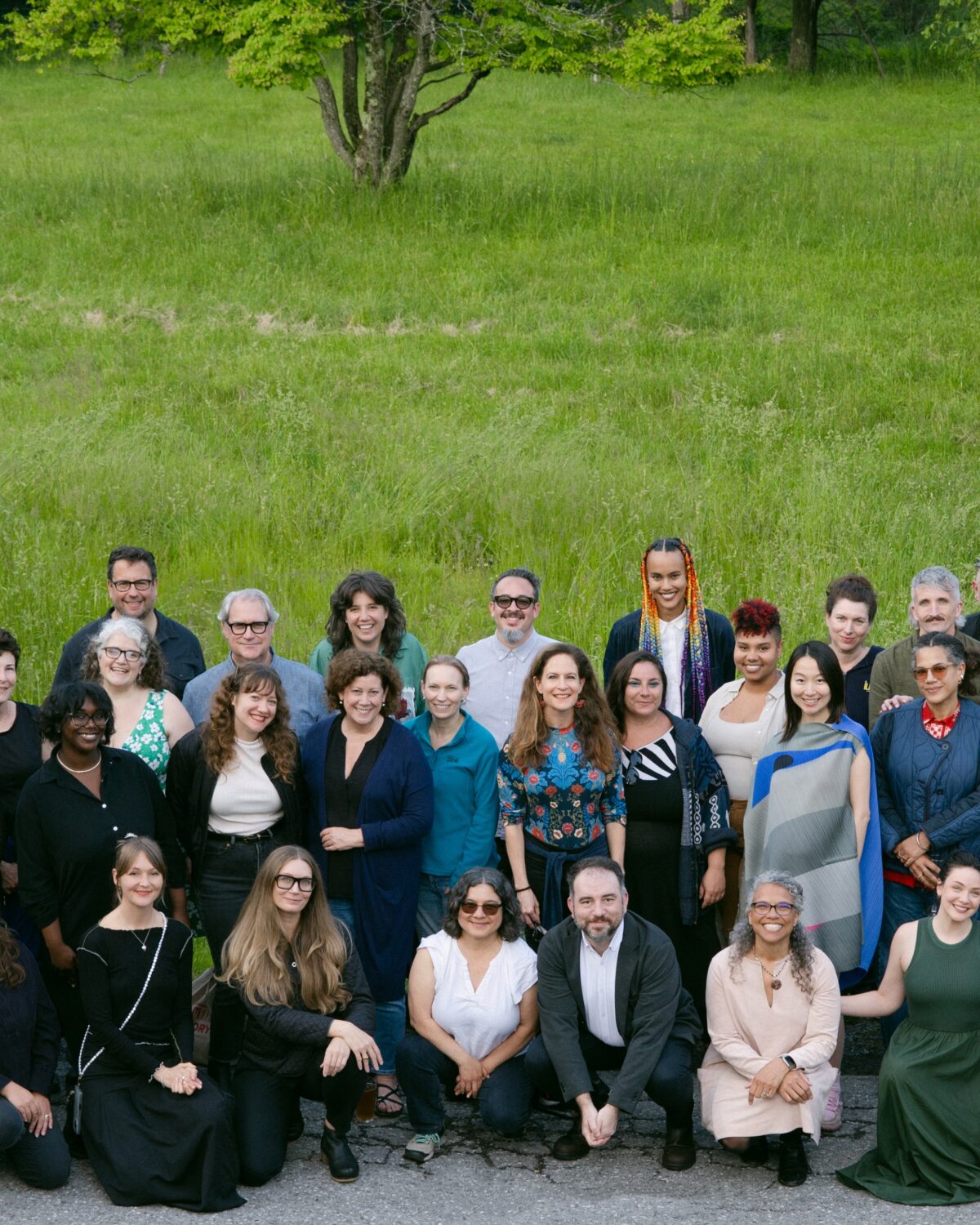 A large group of people pose together outdoors on a paved area in front of green grass and trees, smiling at the camera.