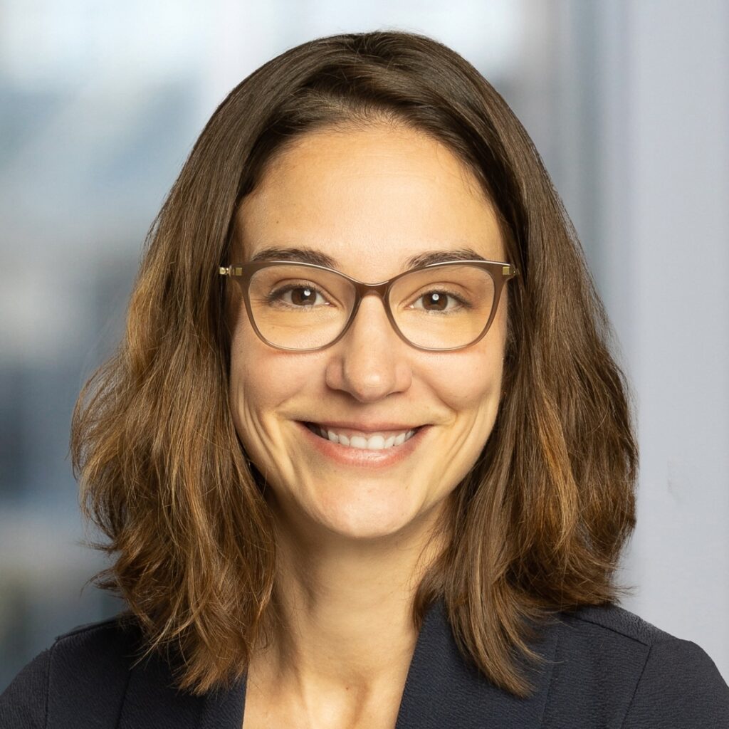 A woman with shoulder-length brown hair, wearing glasses and a dark blazer, smiles at the camera in a brightly lit indoor setting with a blurred background. Photo of Rachel Ward