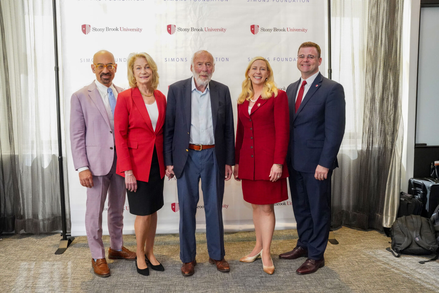 Five people stand smiling in front of a Stony Brook University and Simons Foundation backdrop, dressed in business attire, with two women wearing red and three men in suits.