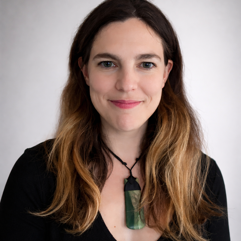 A woman with long brown hair wearing a black top and a green pendant necklace smiles softly at the camera against a plain, light background.