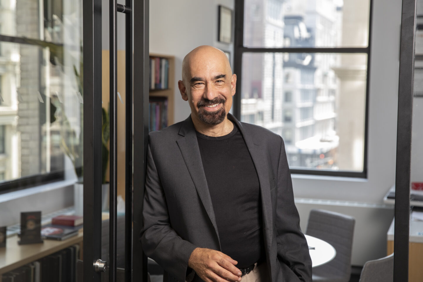 A man with a bald head and goatee, wearing a black blazer and shirt, stands smiling in a modern office with large windows and city views in the background.