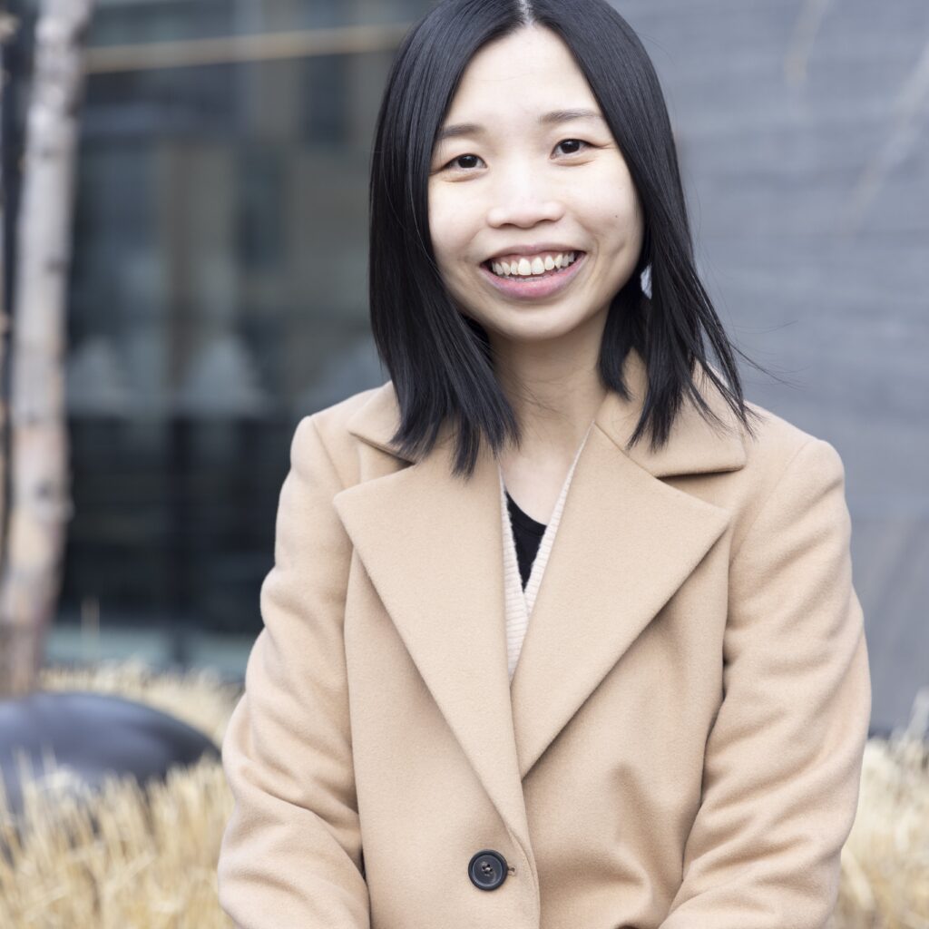 A woman with straight black hair, wearing a beige coat, sits outdoors and smiles at the camera. The background includes blurred buildings and dried plants.