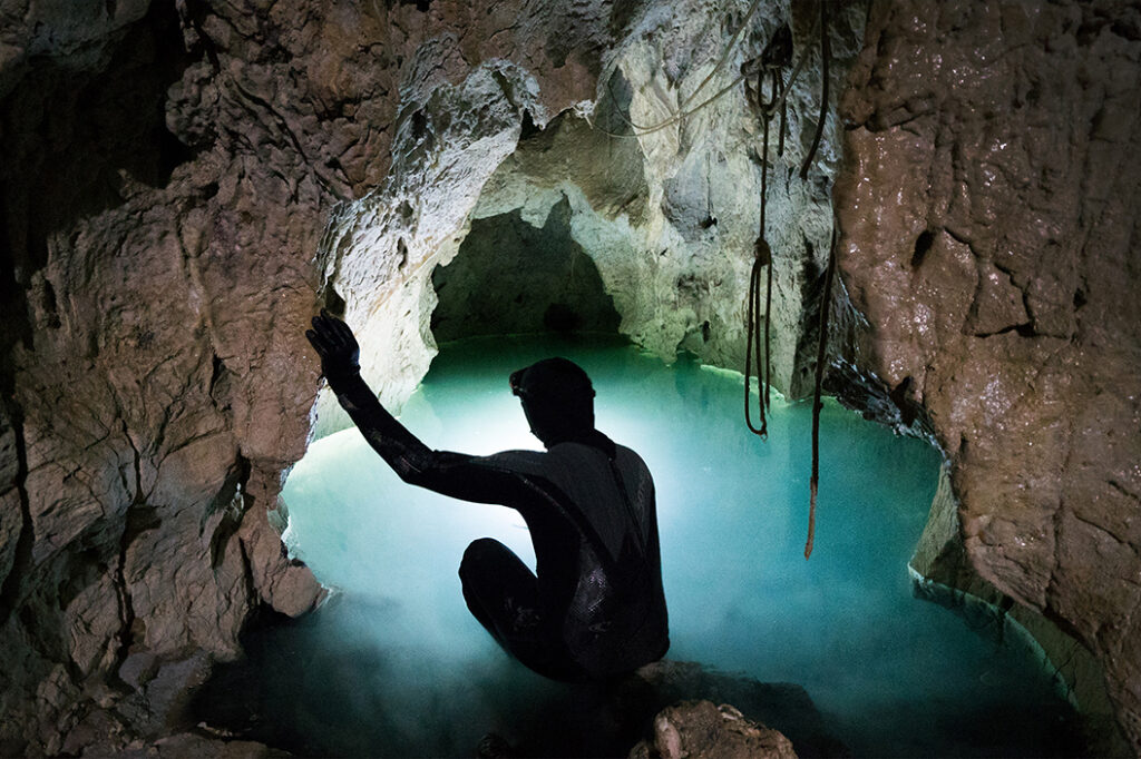 A person in a black wetsuit crouches at the edge of a glowing turquoise underground pool inside a rocky cave, with one hand touching the cave wall.