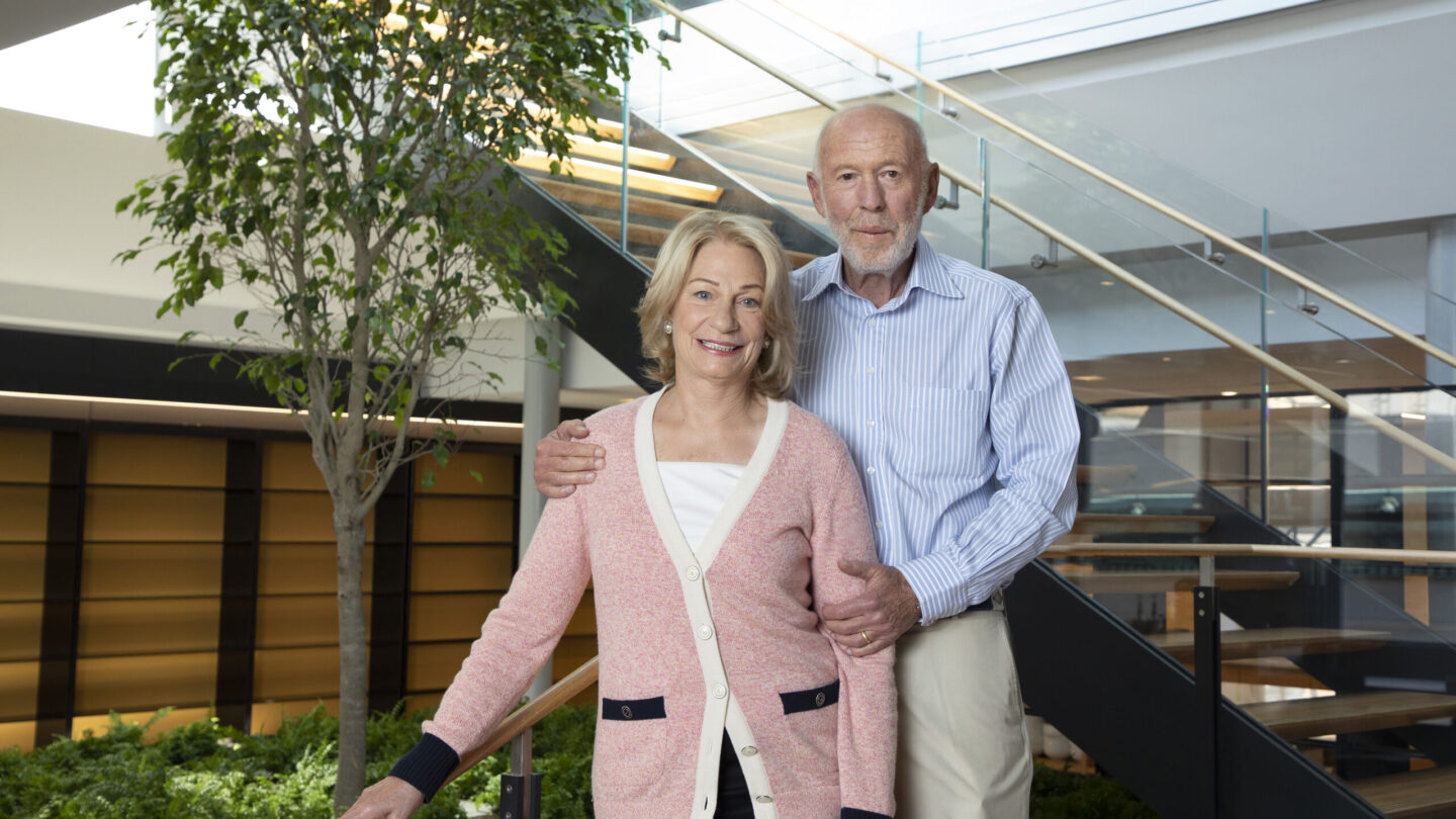 An older couple stands together indoors by a staircase and a tree. The woman wears a pink cardigan and smiles, while the man, in a blue shirt, stands close behind her with his arm around her shoulders.