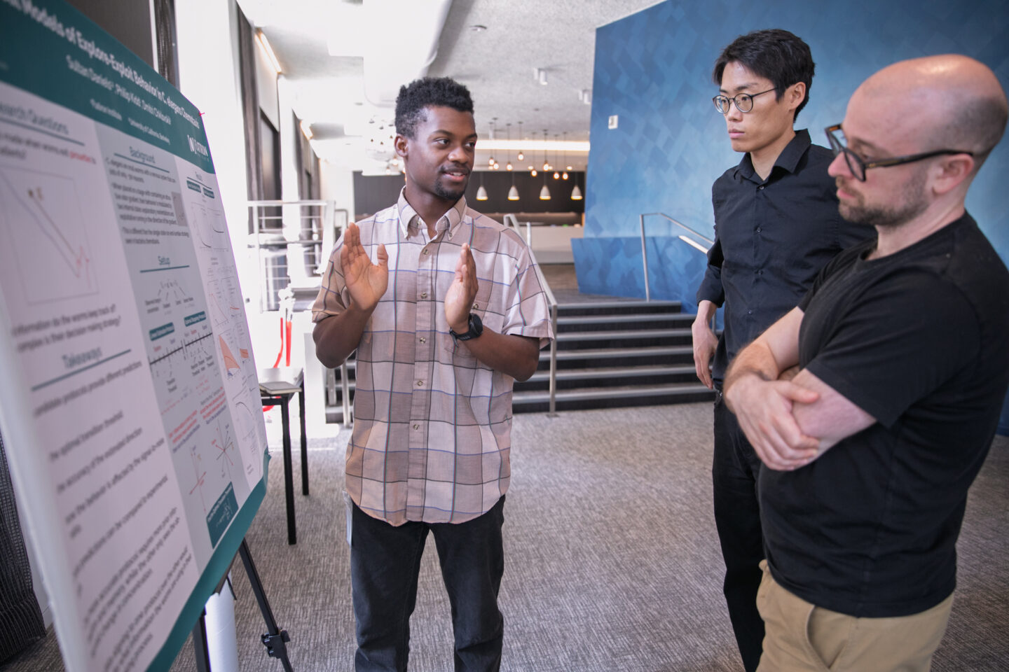 A person explains a scientific poster to two others indoors; one listens with arms crossed, another stands attentively. They are in a modern, well-lit hallway with stairs and geometric wall patterns in the background.