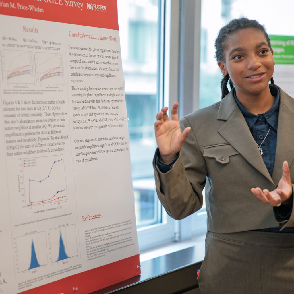 A person in a gray suit gestures while presenting research displayed on a large poster with charts, graphs, and text in a bright, indoor setting.