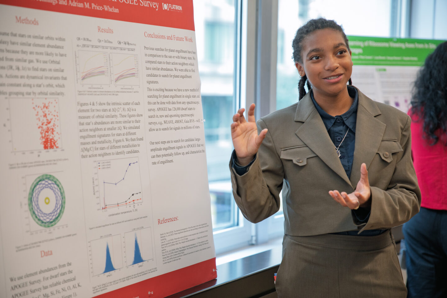 A person in a gray suit gestures while presenting research displayed on a large poster with charts, graphs, and text in a bright, indoor setting.