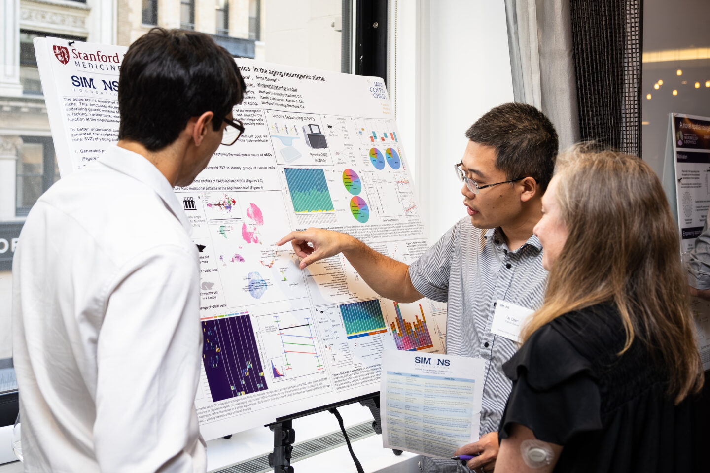 Three people discuss a scientific research poster filled with charts and graphs at a conference. One man points to data, while the others listen attentively. The poster is from Stanford Medicine.