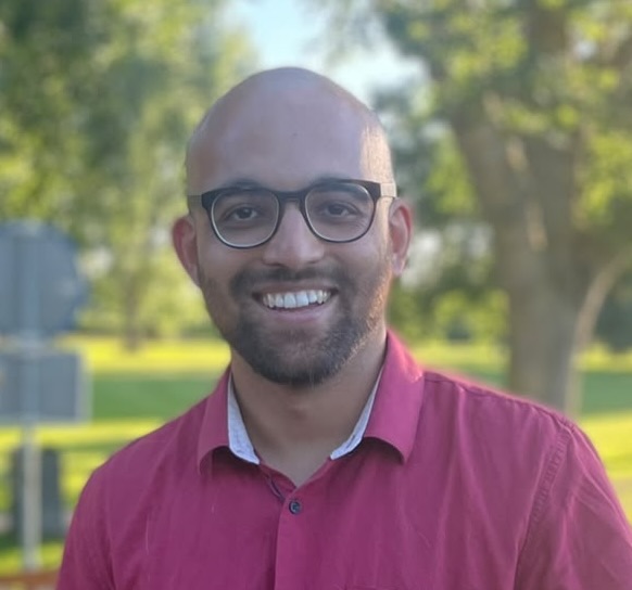 A smiling person wearing glasses and a red shirt stands outdoors in a sunny park with green trees and grass in the background.