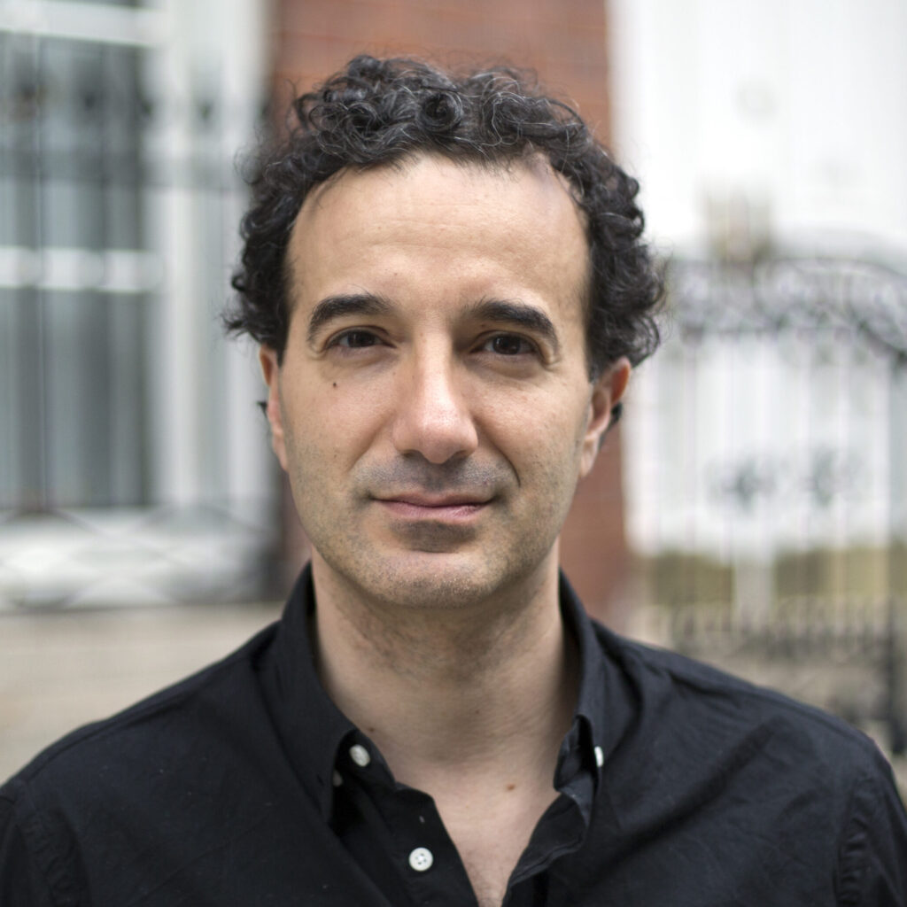 A man with short, curly dark hair wearing a black button-up shirt stands outside in front of a blurred metal fence and brick building, looking calmly at the camera.
