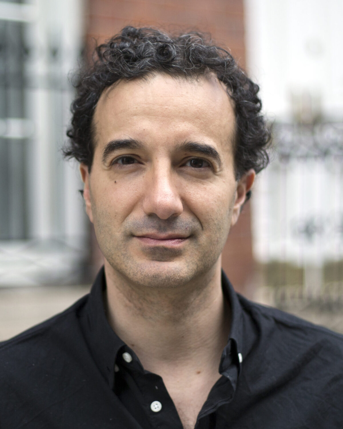 A man with short, curly dark hair wearing a black button-up shirt stands outside in front of a blurred metal fence and brick building, looking calmly at the camera.