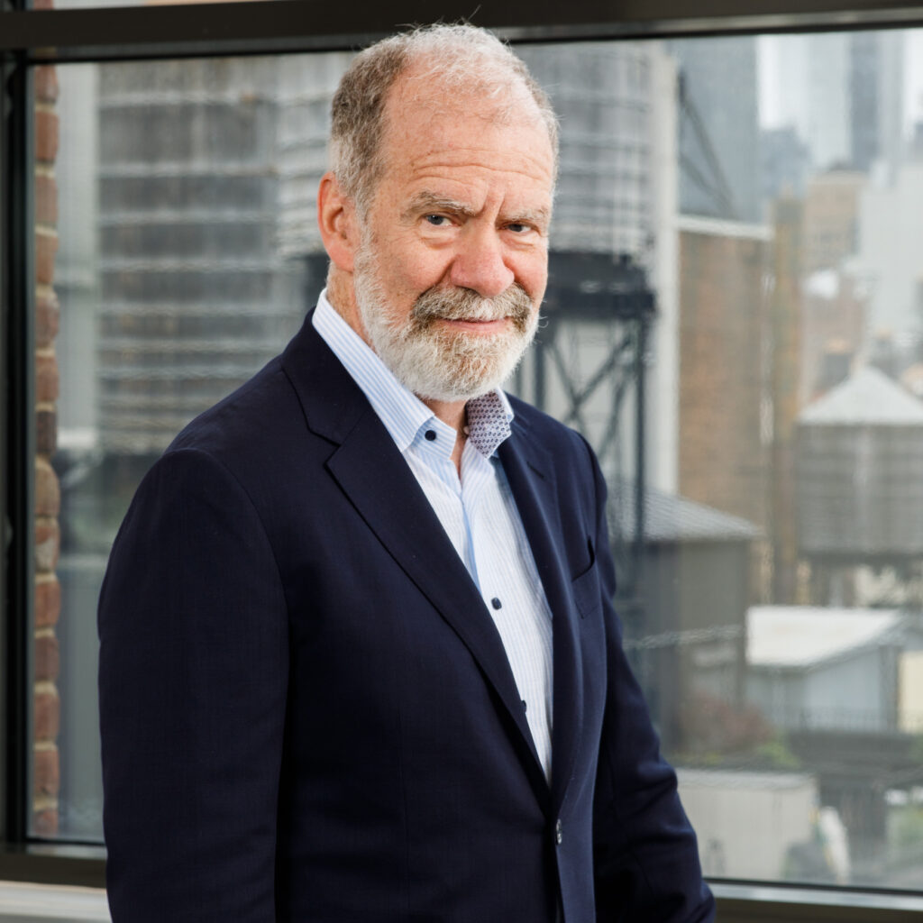 An older man with a gray beard and mustache, wearing a navy suit and light blue shirt, stands in front of a window overlooking an urban cityscape.