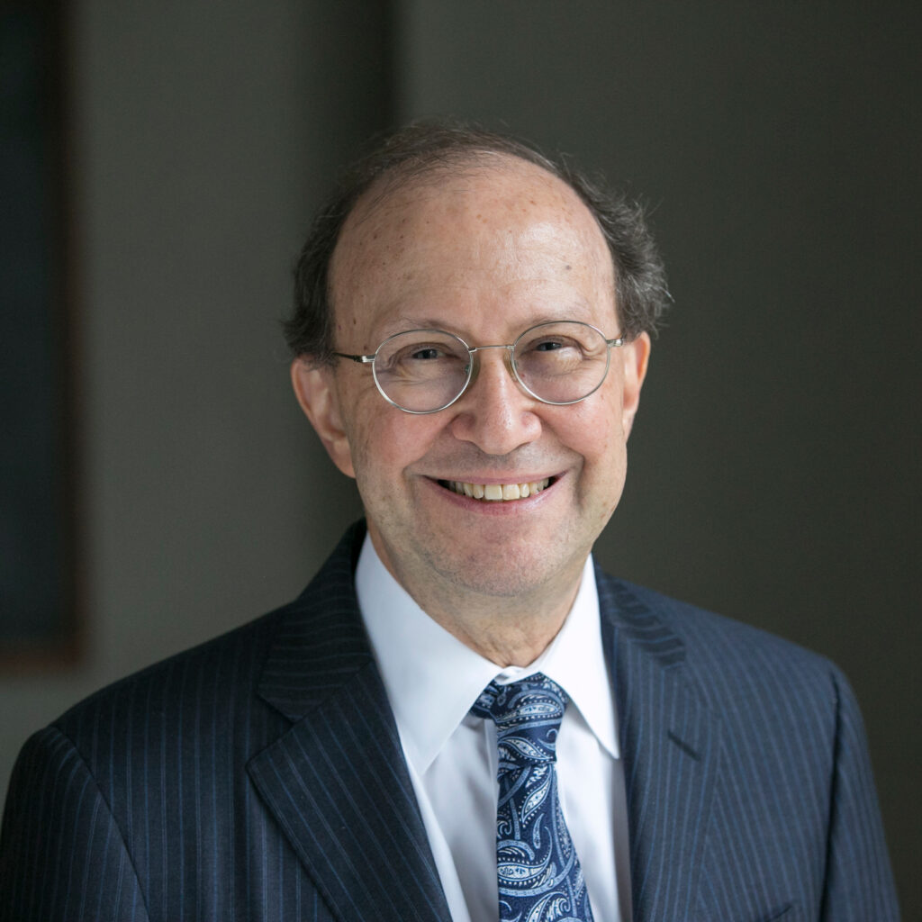 Smiling older man with glasses, wearing a dark pinstripe suit, white shirt, and blue paisley tie, stands in front of a plain dark background.