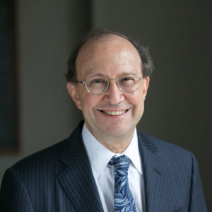 Smiling older man with glasses, wearing a dark pinstripe suit, white shirt, and blue paisley tie, stands in front of a plain dark background.