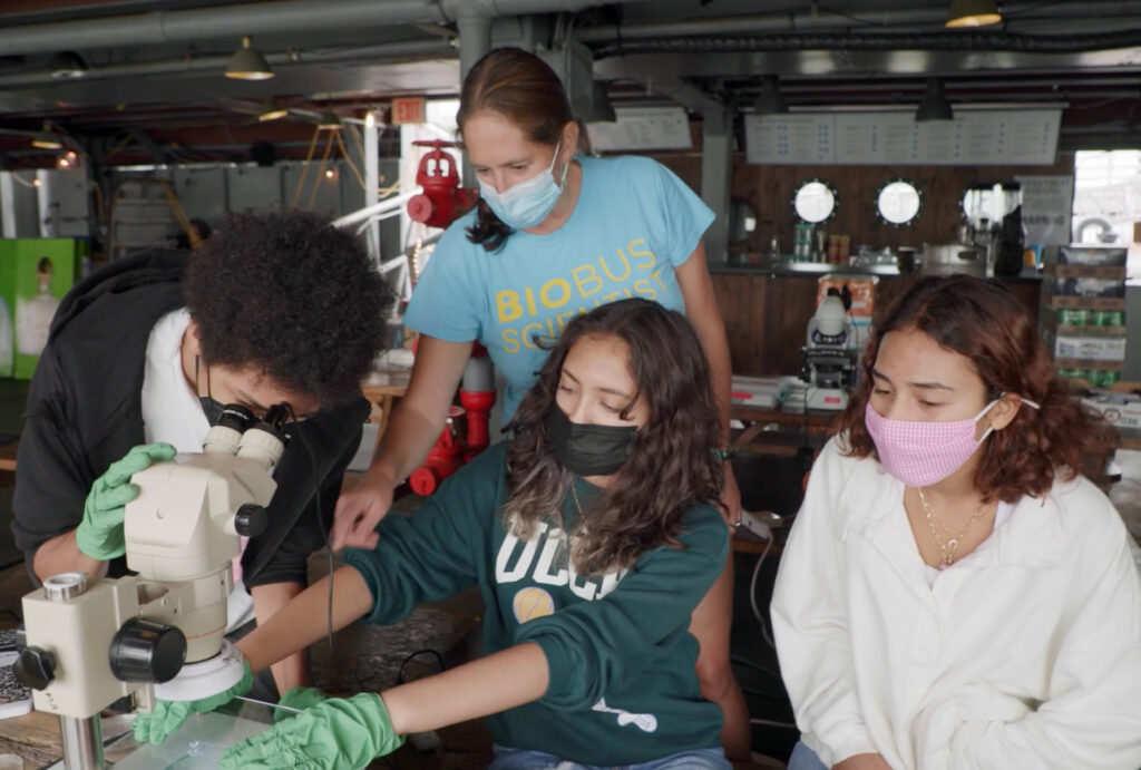 Four students, three wearing masks, work together in a lab. One uses a microscope as another adjusts equipment. An instructor in a BioBus Scientist shirt assists, while another student watches.