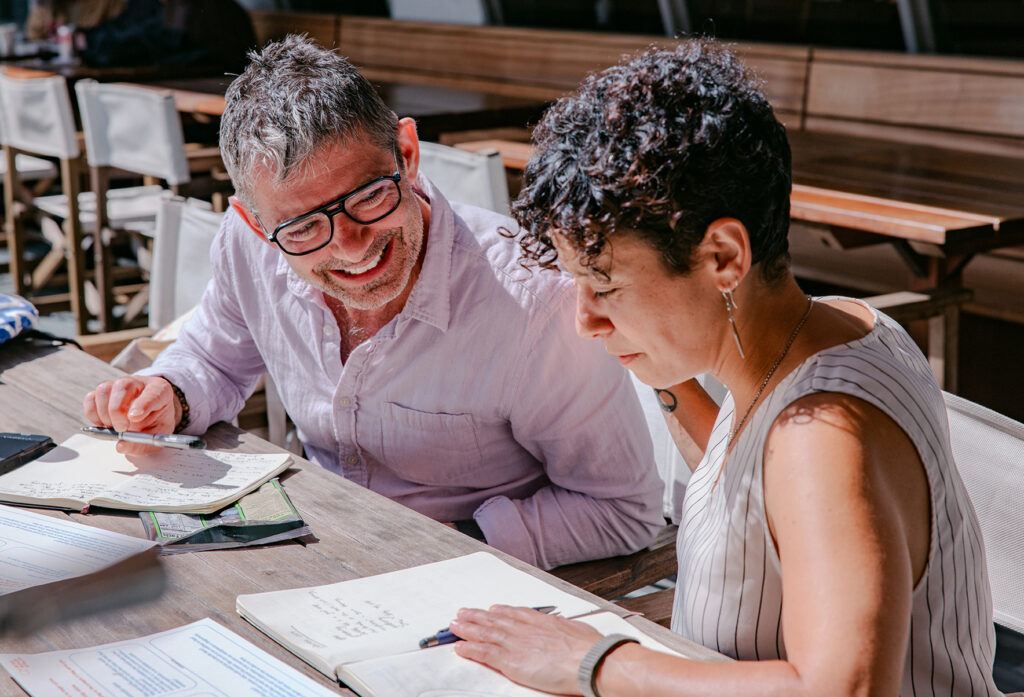 Two people sit at a table, smiling and working together with notebooks and papers. One person writes while the other looks on, engaging in a collaborative discussion in a bright, indoor setting.
