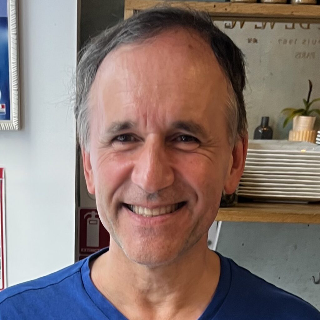 A smiling middle-aged man with short, graying hair wears a blue shirt and stands indoors with shelves, plates, and plants visible in the background.